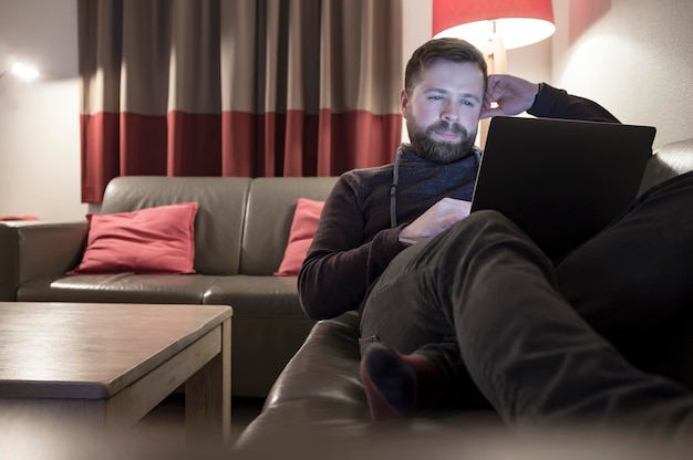 Cute smiling young guy looks into his laptop sitting relaxed on a leather sofa in a comfortable