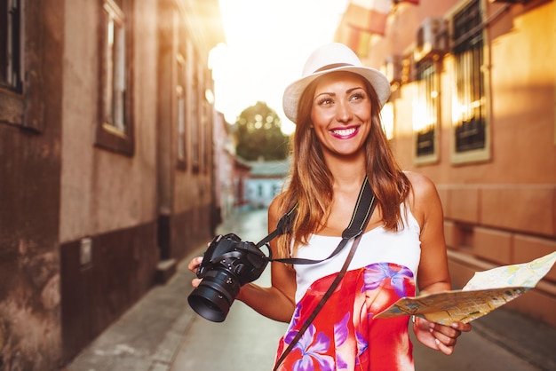 Cute smiling young woman on vacation is walking on the city street. She is holding a digital camera and a city map.