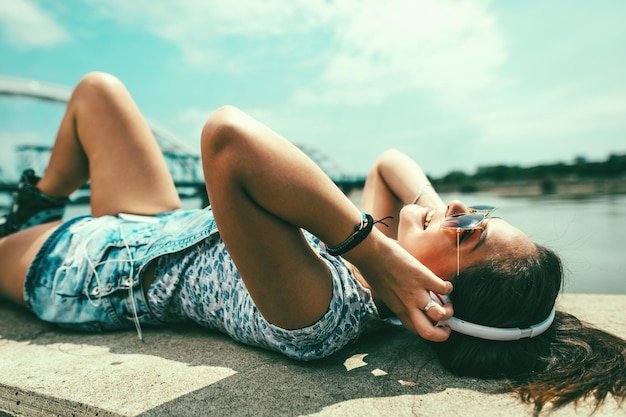 Симпатичная улыбающаяся молодая женщина с роликовыми коньками слушает музыку с телефона и наслаждается лежанием на берегу реки в прекрасный летний день.