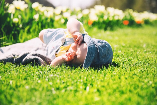 Cute squinting in the sun baby girl lying in green grass of tulip field Child playing outdoors in spring park Image of Mothers Day Easter Family on nature in Arboretum Slovenia Europe