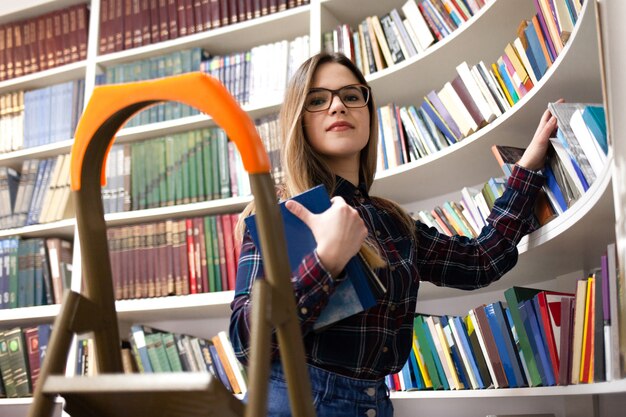 メガネをかけたかわいい学生が図書館の本棚から本を受け取ります。