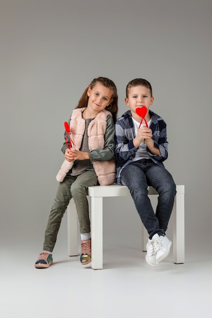 Cute stylish little couple child girl and boy with red hearts on stick in fashionable clothes sittting together