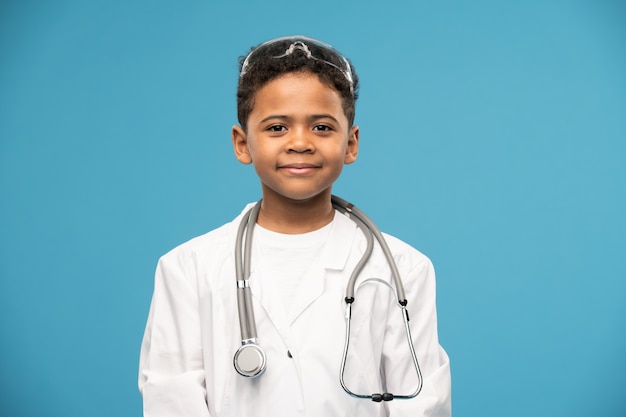 Cute successful little boy of African ethnicity in whitecoat having protective medical eyeglasses on head and stethoscope on neck