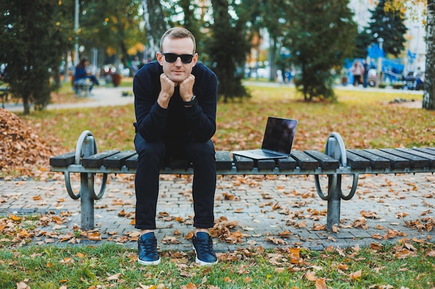 Cute successful young man in sunglasses sitting on park bench\
with laptop and phone and looking away on sunny day freelance\
remote work concept