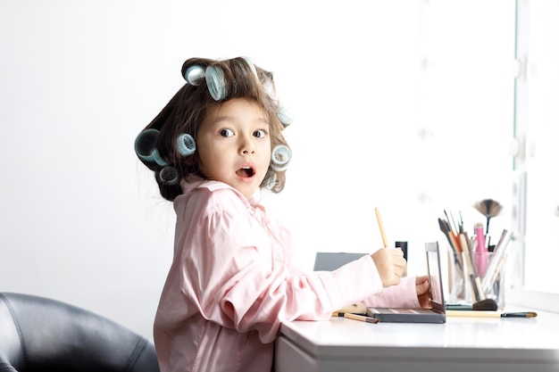 Cute surprised little girl playing with her mother's cosmetic in front of the mirror at home