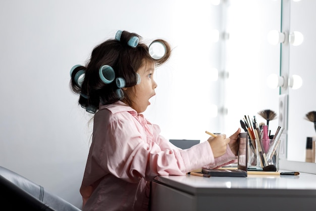 Cute surprised little girl playing with her mother's cosmetic in front of the mirror at home