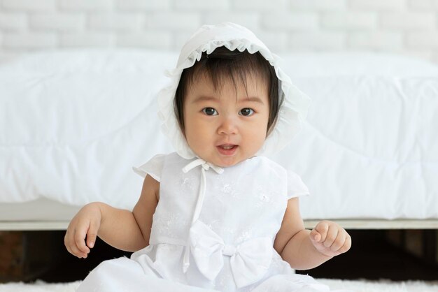 Photo cute sweet adorable asian baby wearing white dress sitting on carpet smiling and playing with happiness emotional in cozy bedroomhealthy baby concept