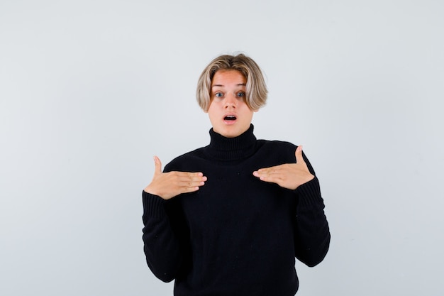 Cute teen boy in black turtleneck sweater with hands on chest and looking shocked