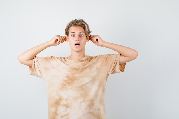 Cute teen boy pulling ears with fingers in t-shirt and looking shocked , front view.