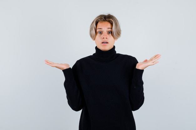 Cute teen boy showing helpless gesture in black turtleneck sweater and looking puzzled , front view.