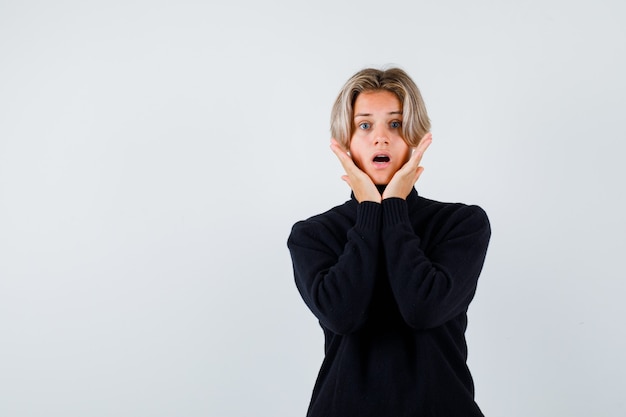 Cute teen boy in turtleneck sweater keeping hands under chin and looking shocked