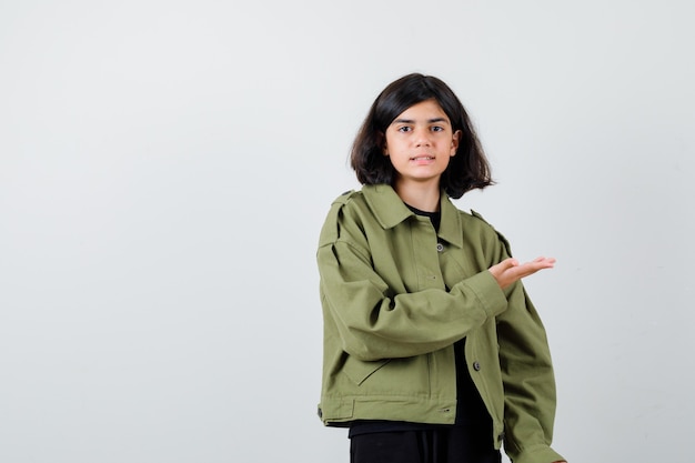 Cute teen girl showing welcoming gesture in army green jacket and looking confident. front view.