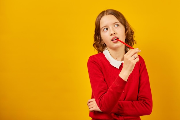 Cute teen school girl is thoughtful with red pencil in hand on yellow studio background back to school concept have an idea copy space