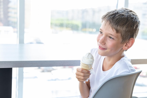 A cute teenage boy eats a swirling white ice cream in a waffle cup sitting at a table by the window in a cafe a copy of the space Blurred background A child enjoys ice cream A sweet snack