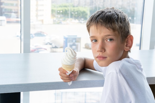 A cute teenage boy eats a swirling white ice cream in a waffle cup sitting at a table by the window in a cafe