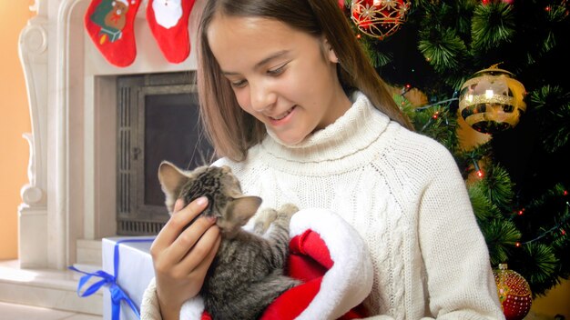 Cute teenage girl caressing grey kitten under Christmas tree
