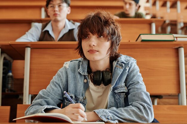 先生の話を聞きながらノートを作るデニム ジャケットでかわいい 10 代の少女