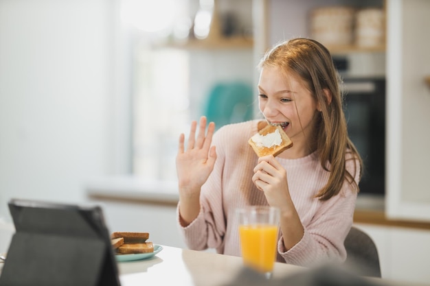 A cute teenage girl having breakfast and making a video call on digital tablet while enjoying suny morning in her kitchen.