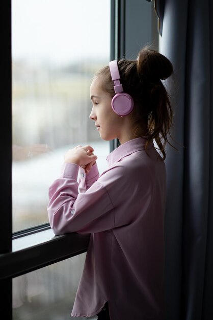 Милая девочка-подросток в розовой рубашке и наушниках смотрит в окно и мечтает