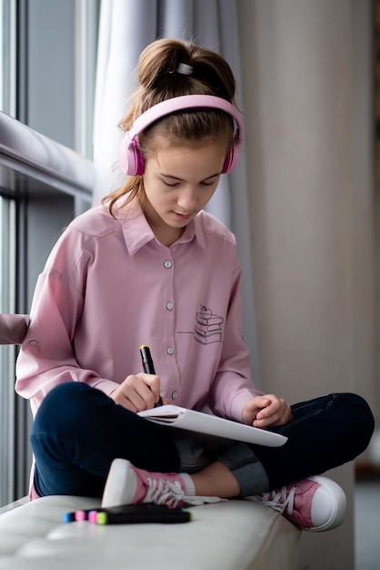 분홍색 셔츠와 헤드폰을 입은 귀여운 10대 소녀가 창 근처에 앉아 마커로 그림을 그립니다.