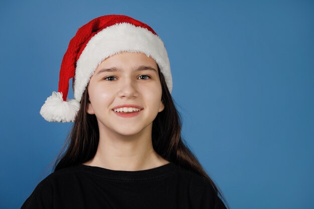 青い背景のカメラでポーズをとって、赤いサンタキャップのかわいい10代の少女が微笑む。