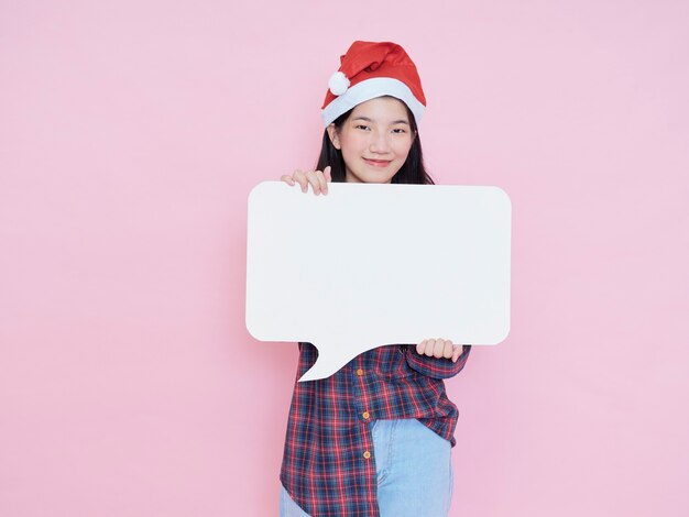 ピンクの背景に白い空白のポスターを保持しているサンタの帽子のかわいい10代の少女