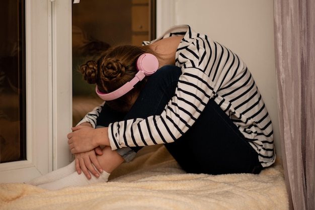 Photo a cute teenage girl sits on a window listening to music with headphones and thinking