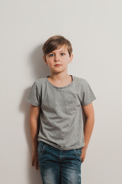 cute teenager boy stands next to white wall, grey T-shirt, blue jeans, frightened face