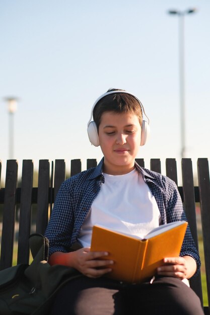 a cute teenager boy with headphone reads yellow book sits on bench outdoor in the park on blue sky