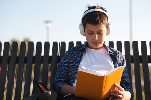 A cute teenager boy with headphone reads yellow book sits on bench outdoor in the park on blue sky