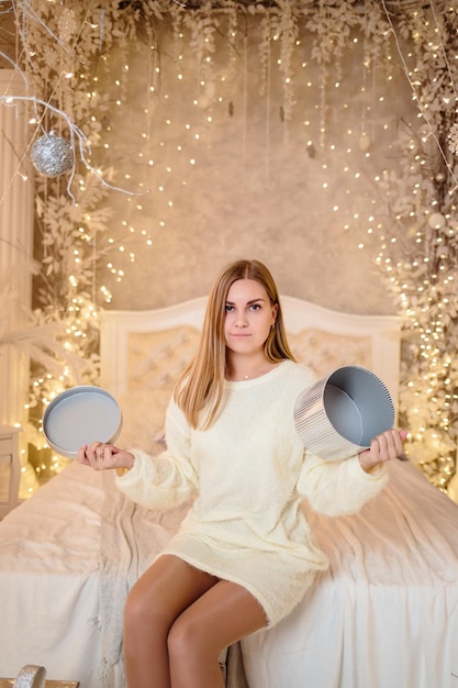 A cute tender beautiful woman sitting on a white bed opens a wrapped gift on Christmas morning New Years celebration home decoration with lights and trees