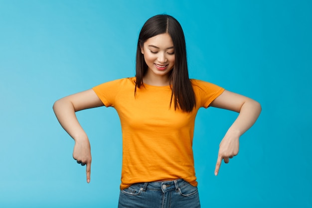 Cute tender feminine asian girl dark haircut look pointing down contemplating interesting product, stare curiously smiling amused, express enthusiasm interest try-out cool thing, blue background.