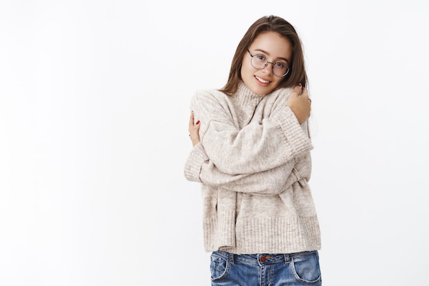 Cute and tender womanfriend in glasses hugging herself and leaning head on shoulder as feeling lonely, missing boyfriend on cold winter day, wearing warm sweater to feel comfort over gray wall.