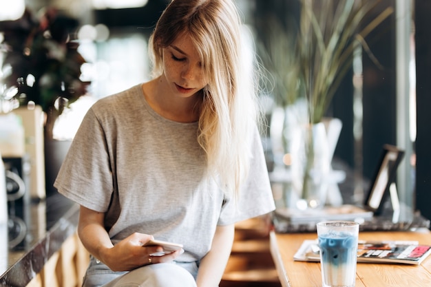 A cute thin blonde girl,dressed in casual style,drinks coffee and looks at her phone in a coffee shop. .