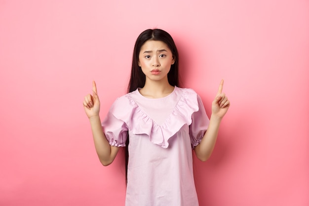 かわいい臆病なアジアの女性は、指を上に向け、眉をひそめ、動揺し、ロゴに指を向け、ピンクの背景にドレスを着て立っています。