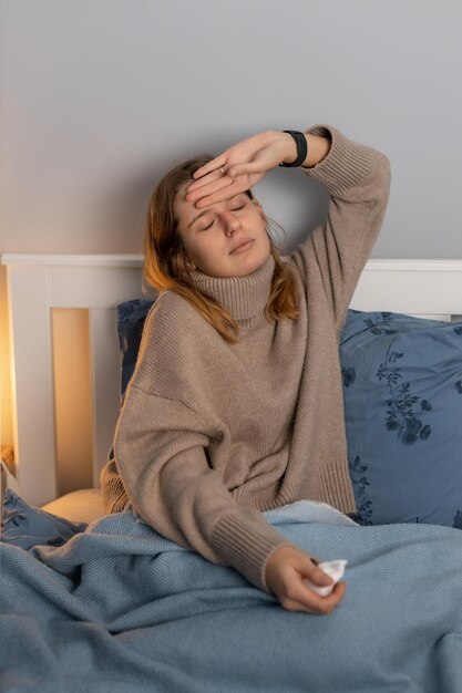 Cute tired caucasian girl in beige sweater sitting on bed and holding her head. Sickness woman touching her forehead, suffering from heat. Female got the flu or a cold. Healthcare concept.