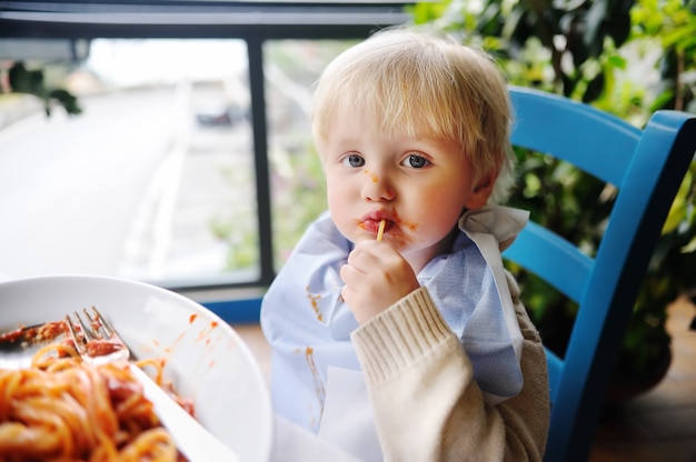 Cute toddler boy eating pasta in Italian indoors restaurant. Healthy/unhealthy food for little kids