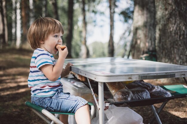 Cute toddler boy eating piece of bread outdoors Summer picnic in nature Portrait of joyful child sitting on camping chair and holding in his hands brioche in forest