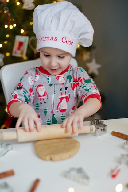 Cute toddler boy making Christmas cookies on a white table near Christmas tree with lights Christmas cooking concept