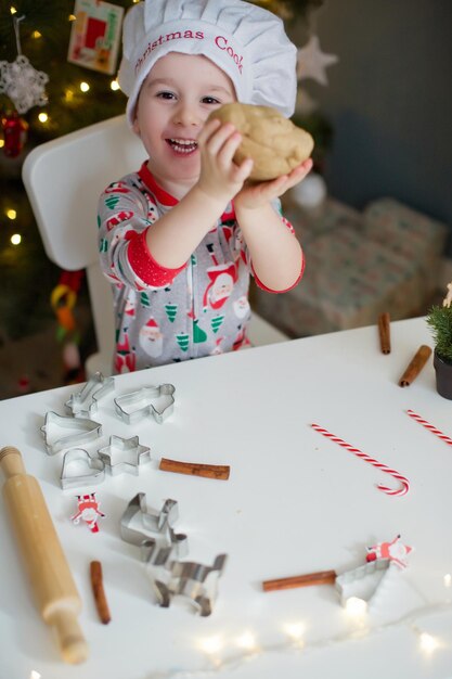 크리스마스 요리 컨셉으로 조명이 있는 크리스마스 트리 근처 흰색 테이블에서 크리스마스 쿠키를 만드는 귀여운 소년