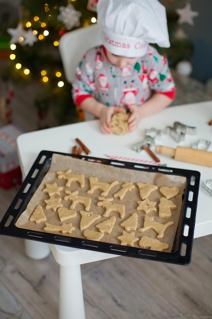 ライトとクリスマスツリーの近くの白いテーブルでクリスマスクッキーを作るかわいい幼児の男の子クリスマス料理のコンセプト