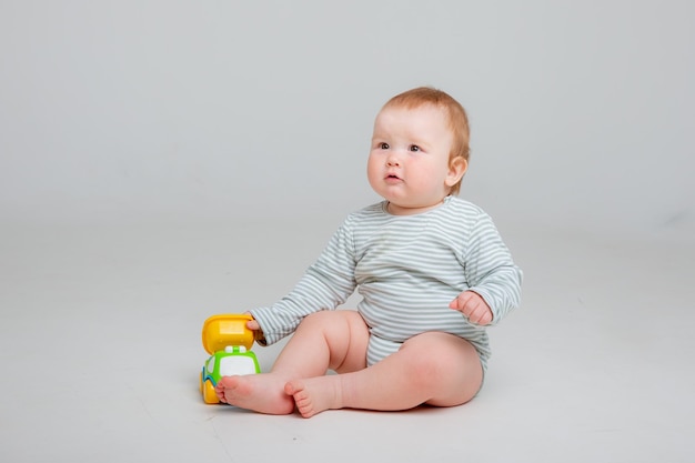 Photo cute toddler boy old in white bodysuit plays with car toy sitting on a white background space for te