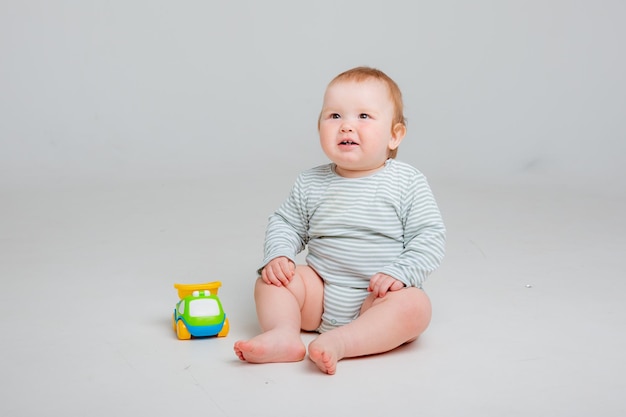 Cute toddler boy old in white bodysuit plays with car toy sitting on a white background space for te