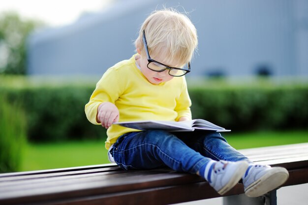 Cute toddler boy reading a book outdoors on warm summer day. Back to school concept