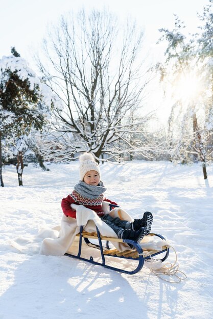 晴れた冬の日に雪に覆われた都市公園のそりに座っているかわいい幼児の男の子