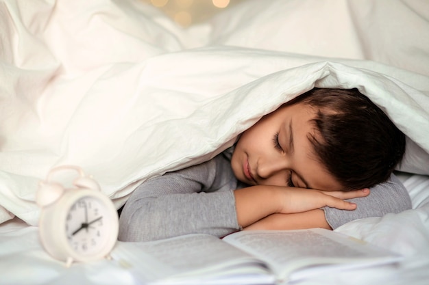 Cute toddler boy sleeping with a book under a white blanket.