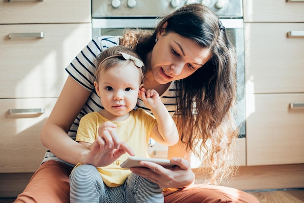 Cute toddler girl touching her ear and smiling while sitting with her mother on the kitchen floor stock photo