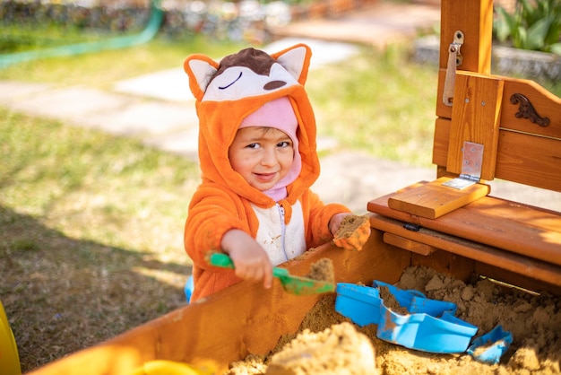 オレンジ色のジャンプスーツを着たかわいい幼児は、晴れた日に屋外のサンドボックスの砂で遊ぶ