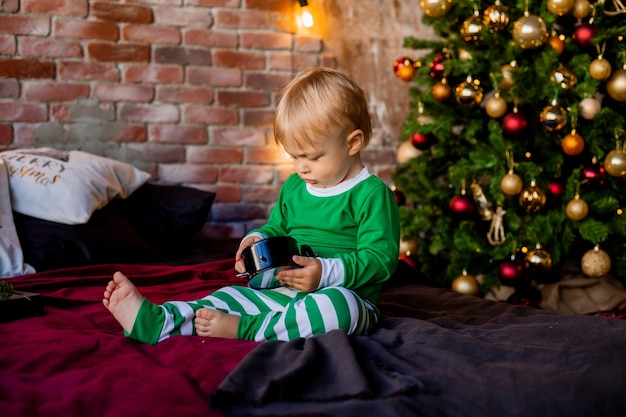 Cute toddler in pajamas sits near Christmas tree