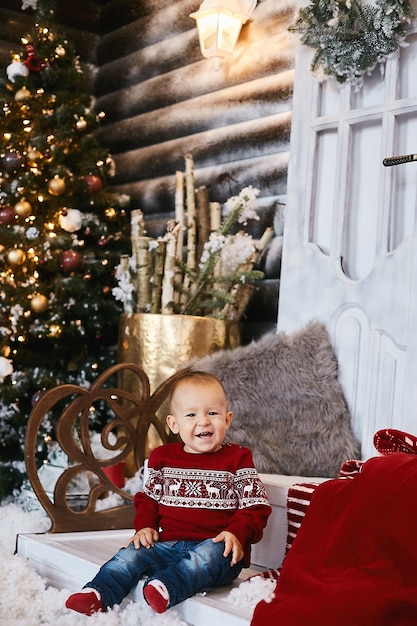 크리스마스 집으로 장식된 계단에 포즈를 취한 못생긴 스웨터를 입은 귀여운 유아